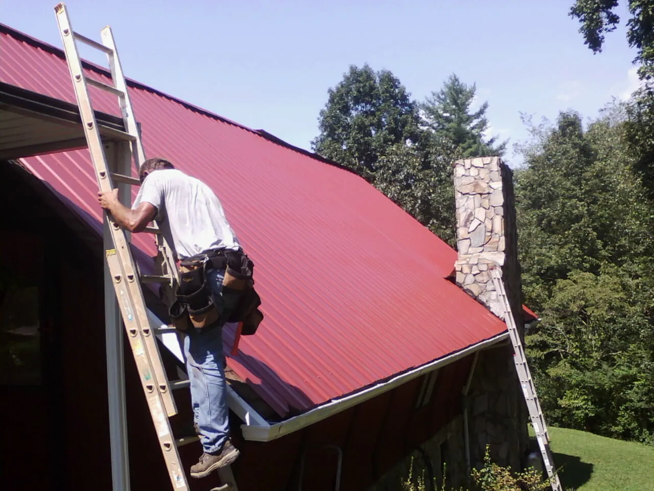 Roofer on a ladder beside a red metal roof and stone chimney — Soffit & Fascia Repair by Roofing Brothers in Leota